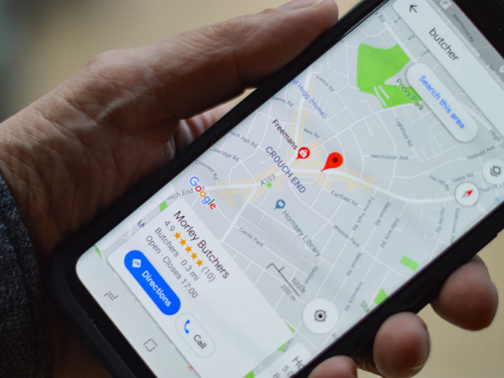 A hand holding a phone showing a Google Maps search result for a local butcher shop with a five-star rating and call and directions buttons
